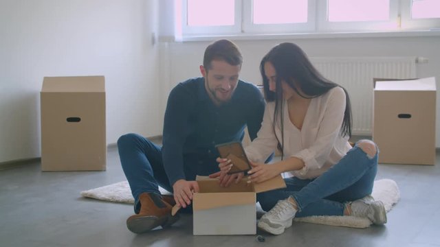 Young caucasian couple sitting on floor of new house unboxing the photos getting happy and excited.