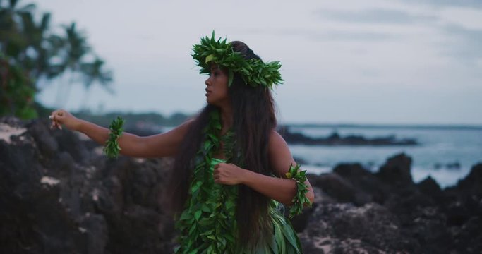 Traditional Hawaiian hula dancing at sunset in slow motion, woman performing Hawaiian hula with haku leis and ti leaf skirt with the ocean in the background