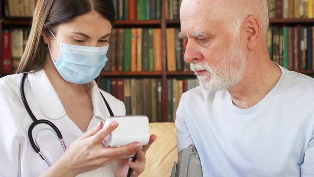 Female professional doctor in medical mask with stethoscope at work. Woman physician measuring pressure to sick senior male patient by tonometer at home. Consulting about treatment and therapy
