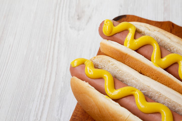 Two hot-dogs with yellow mustard on a rustic wooden board on  a white wooden background, low angle...