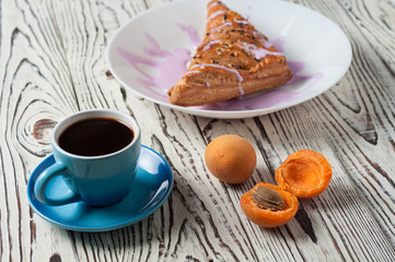 Full ceramic cup of hot black coffee near baked sweet bun in form of triangle with purple cream and apricot lies on white weathered wooden table