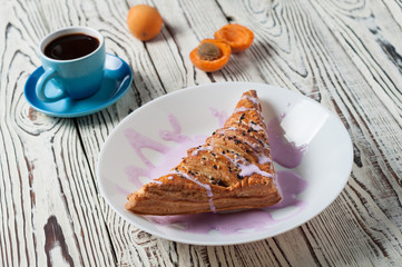 Full ceramic cup of hot black coffee near baked sweet bun in form of triangle with purple cream and apricot lies on white weathered wooden table