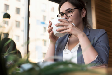 Beautiful brunette business woman in white skirt and grey suit jacket working from the cafe. She wears glasses and drink a cup of coffee. copy space