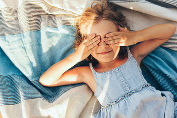 Beautiful blonde little girl lying on the blanket, cover her eyes from the sun with both hands,...