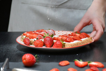 An image of a plate with two strawberry sandwiches in a girl’s hand; powdered sugar snowing down on sandwiches, a cutting board with sliced & whole strawberries, a knife on a black wooden table