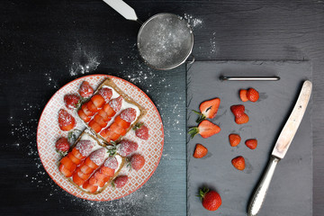 An image of a plate with two strawberry sandwiches, a cutting board with sliced and whole strawberries, a knife, a small sieve and powdered sugar around on a black wooden table