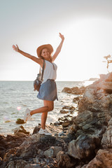 A young hipster woman in a hat and a rukzak with her hands up, standing on top of a cliff and looking at the sea at sunset.