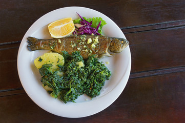 Balkan cuisine. Grilled fish ( trout  ) with leafy green vegetables on white plate. Dark rustic background. Flat lay, free space for text