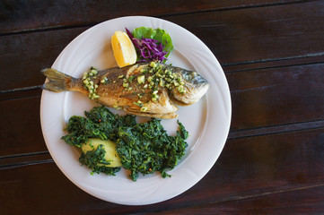 Balkan cuisine. Grilled fish ( sea bream ) with green leafy vegetables on white plate. Dark rustic background, flat lay, free space for text