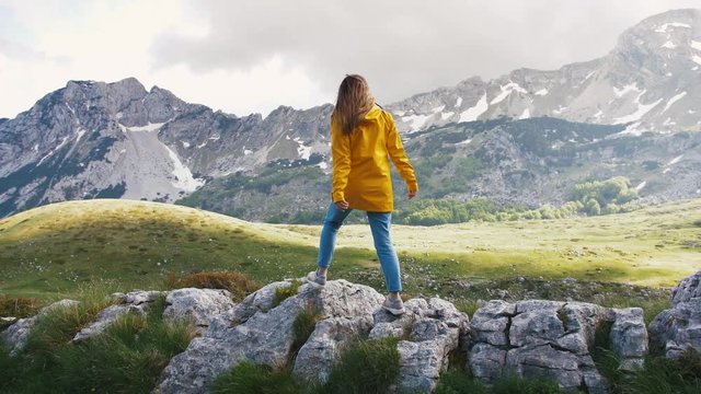 Young cheerful woman in a yellow raincoat walking on rocks and rising hands with beautiful mountains background, slow motion