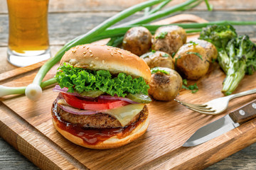 Delicious cheeseburger with baked potatoes, broccoli and scallion on a cutting board served with a glass of lager beer.