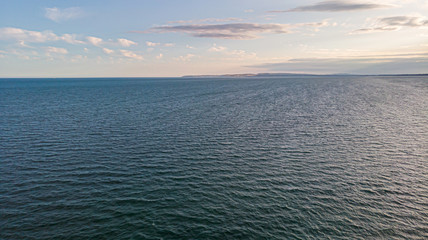 An aerial view of a majestic choppy blue water sea with some hilly coastline in the background under a majestic blue sky and white clouds