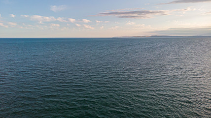 An aerial view of a majestic choppy blue water sea with some hilly coastline in the background under a majestic blue sky and white clouds
