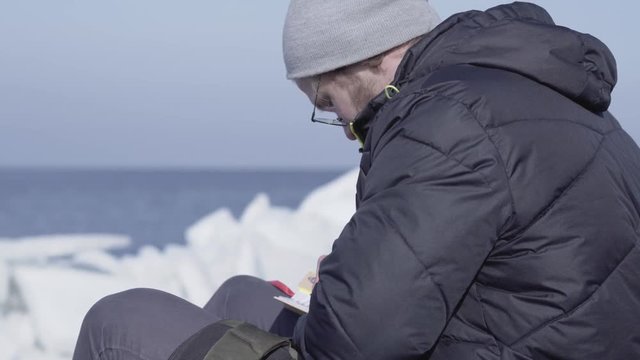 Young man in warm jacket and hat sitting on the glacier writing down his thoughts in a little notebook. Amazing nature of a snowy glacier. The polar explorer on the block of ice