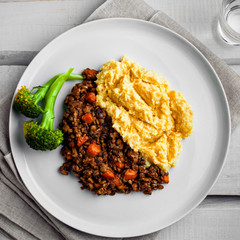Plate of shepherd’s pie on a white wooden table. Meal made of smashed potatoes, minced meat, lentil and vegetables. Top view, above shot.