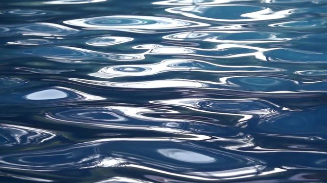 Water Surface Background stock footage features an extreme close-up shot of blue water in a swimming pool. The water is rippling in slow motion and has some reflections of the sun.