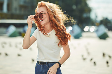 Beautiful red-haired girl having fun on the street. The girls have a beautiful figure, a white T-shirt and jeans with sunglasses - obrazy, fototapety, plakaty