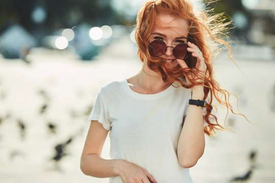 Beautiful red-haired girl having fun on the street. The girls have a beautiful figure, a white T-shirt and jeans with sunglasses