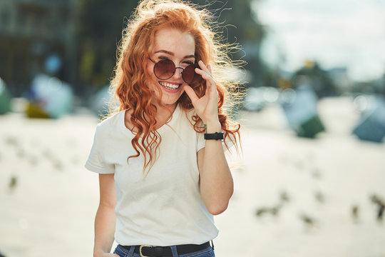 Beautiful red-haired girl having fun on the street. The girls have a beautiful figure, a white T-shirt and jeans with sunglasses