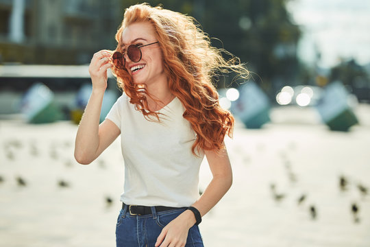 Beautiful red-haired girl having fun on the street. The girls have a beautiful figure, a white T-shirt and jeans with sunglasses