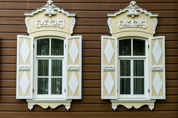 two windows with the wooden carved architrave in the old wooden house in the old Russian town. - obrazy, fototapety, plakaty