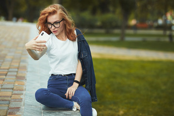 A beautiful red-haired girl is resting in a public garden and takes a selfie on her mobile phone. The girl is wearing a T-shirt with jeans, glasses on her face, and a modern gadget on her arm