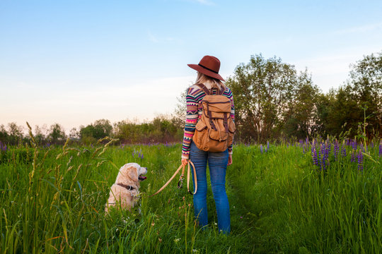 Traveling woman and golden retriever dog in nature. Stylish hipster in brown hat and boho style sweater with vintage textile backpack in summer green meadow. Travel and wanderlust concept.