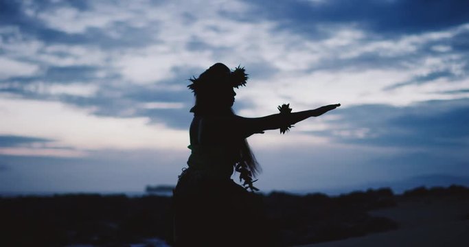 Silhouette of a traditional Hawaiian hula dancer woman dancing on a rugged island landscape at dusk in slow motion with an ocean background