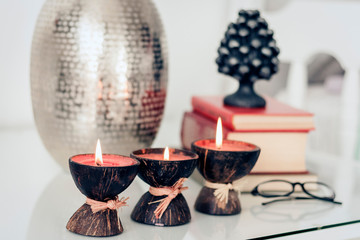 Cozy home interior decor, burning spa aroma candles in coconut shell, metal vase, pile of books, on white glass nightstand.