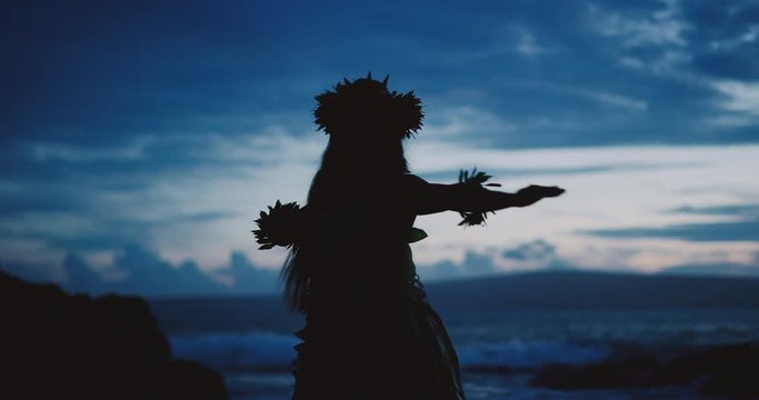 Silhouette of a traditional Hawaiian hula dancer woman dancing on a rugged island landscape at dusk in slow motion with an ocean background
