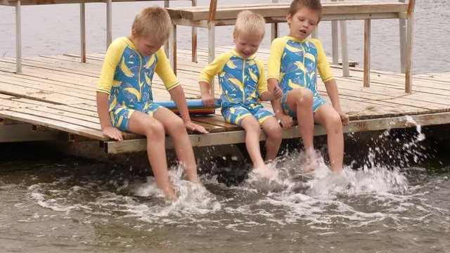 Funny children in bathing suits sit together on wooden bridge, legs in water. boys are happy on holiday in village together. Summer day, river, swimming in water. Closeup of legs and feet