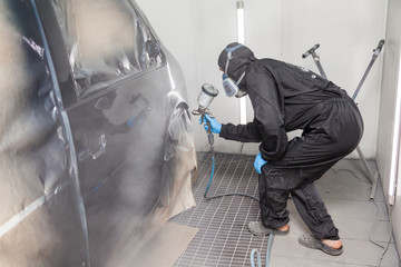 A male worker in jumpsuit and blue gloves paints with a spray gun a side part of the car body in black after being damaged at an accident. Auto service industry professions