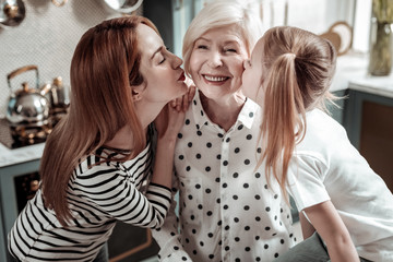 Naklejka premium Aged woman smiling while daughter and granddaughter kissing her