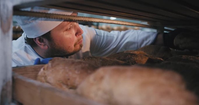 Big bakery man in a uniform inside of industrial shelves take a fresh baked organic bread and smelling deep