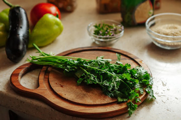 Fresh greenery on the cutting board. Eggplant, pepper and tomato lie on the table in the background.