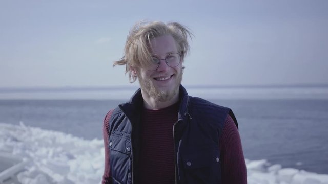 Close-up portrait of attractive young blond man in glasses looking away touching his hair. Attractive guy of Scandinavian appearance in front of winter landscape at the North or South pole
