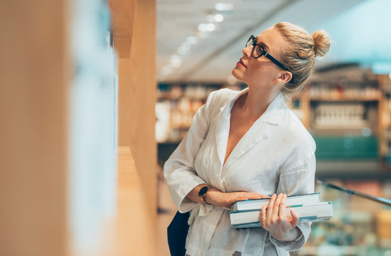 Pretty Caucasian serious woman standing at library and holding books.