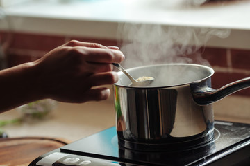 The pot is on the stove in the kitchen. Female hand stirs porridge. Boiling water rises steam.
