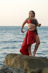 A woman in a red costume for dance with an oriental saber on a belt is posing posing on a rock washed by sea waves.