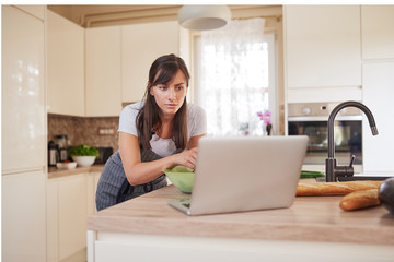 Pretty Caucasian brunette in apron leaning on kitchen counter and looking at laptop and following recipe for dinner.