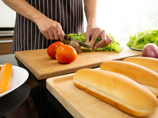 Asian chef man chopping fresh green lettuce root and slicing tomatoes for preparing lunch ingredient delicious hot dog fast food on table in the kitchen