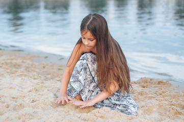 Little girl with long hair in summer dress playing with sand on the beach