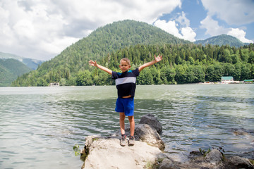 Portrait of a boy with open arms standing on a stone on a lake against a mountain. Lake Rizza Abkhazia.