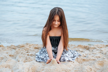 Little girl with long hair in summer dress playing with sand on the beach