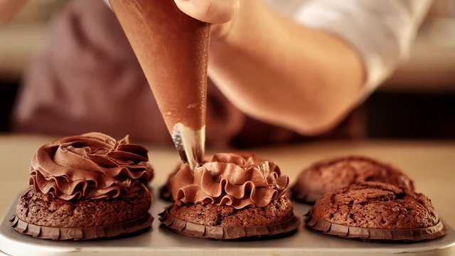 Confectionery decorator. Pastry chef squeezing chocolate frosting on cupcakes from piping bag