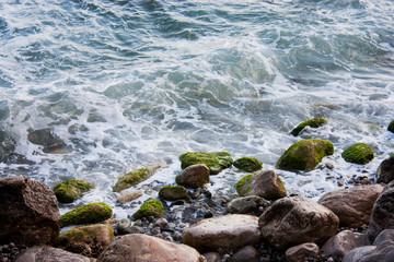 Stone beach of the Black Sea with magnificent waves of the surf. View of the surf waves on the sea beach on a summer day. Foam from the surf on the stones close-up.
