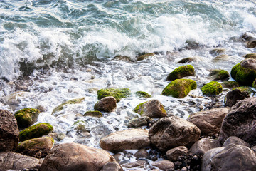 Stone beach of the Black Sea with magnificent waves of the surf. View of the surf waves on the sea beach on a summer day. Foam from the surf on the stones close-up.