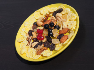 Tasty and beautiful dish is with black and red currant berries, corn flakes, candied fruit  and assorted nuts in a yellow plate on a dark background. Prepare a delicious and healthy dessert for Breakf