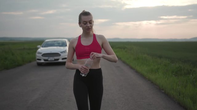 Fitness woman finish run training on green field drink water walk background car runner sport female health outdoor young body digital rural energy fit fitness music player warm close up slow motion