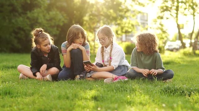 Kids and gadgets. Classmates pass each other a smartphone while sitting on the grass in the park. Sun set